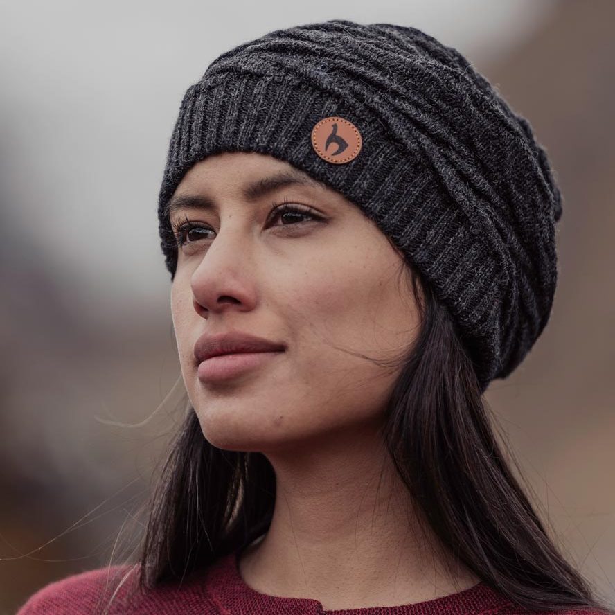 Akaari - Premium Alpaka Mützen Akaari - Premium Alpaka Mützen - Zwei lächelnde Frauen in traditioneller Kleidung stehen neben einem Lama vor bewölktem Himmel.